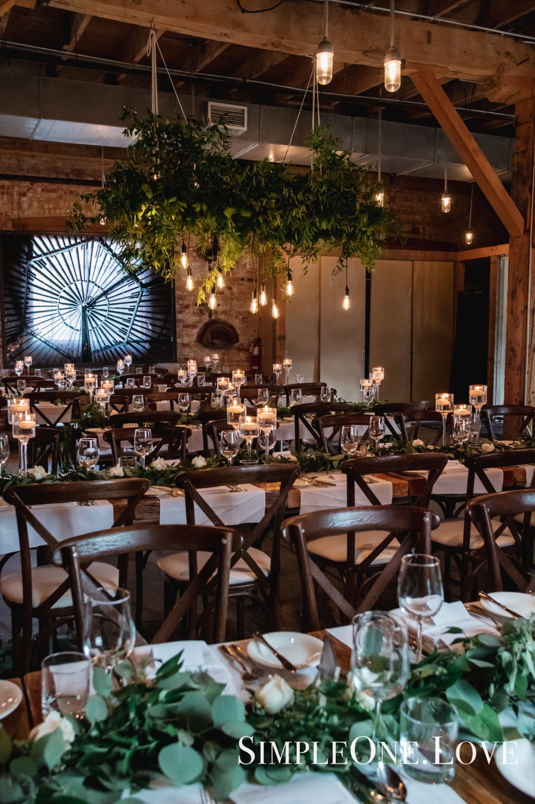 Long wooden tables under greenery in a brick-and-beam room.