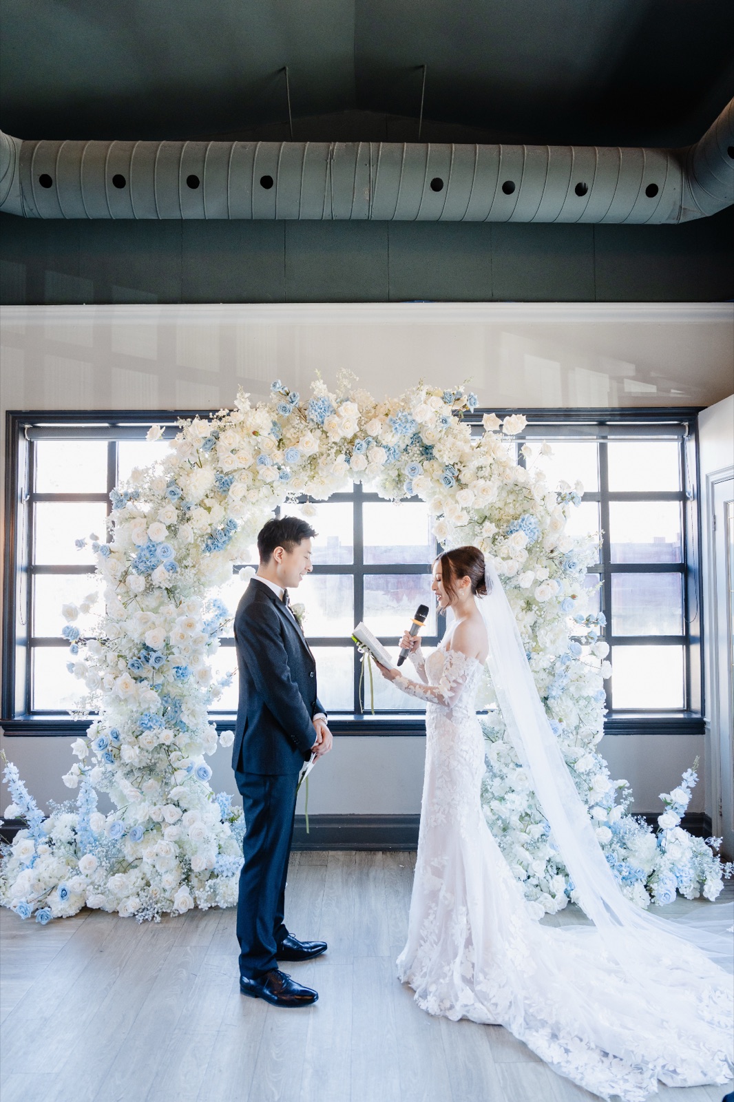 A couple under a white arch in a room of tall windows.