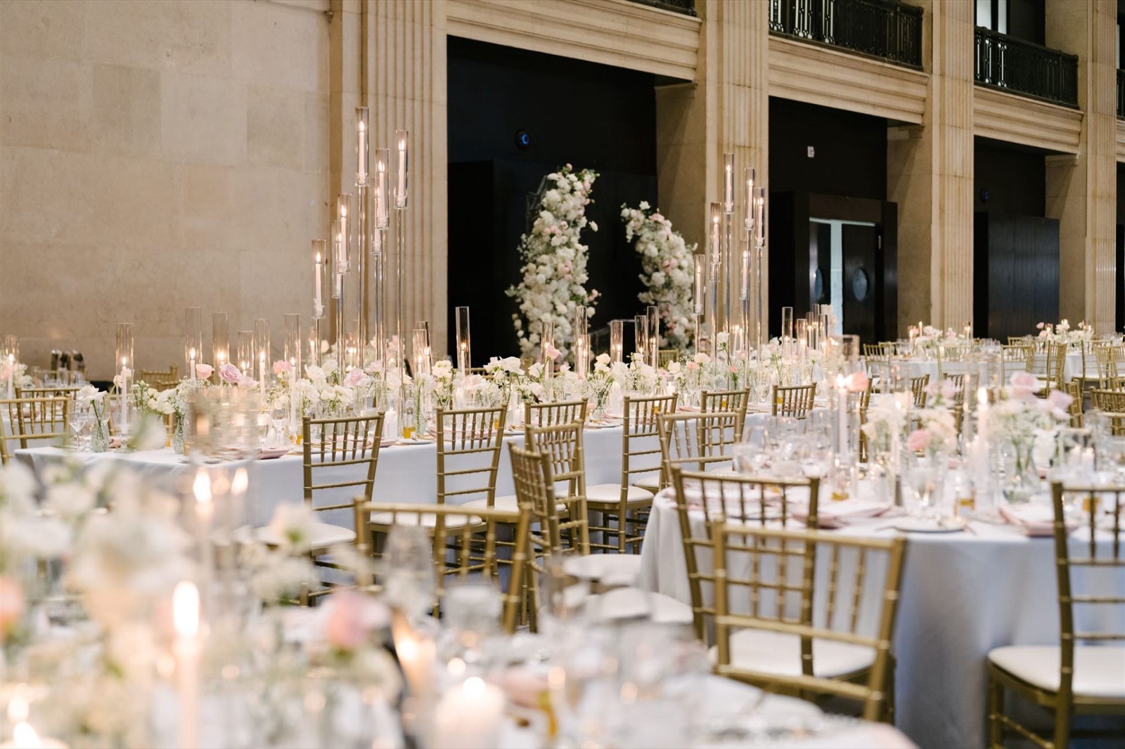 Round tables and gold chairs set for a reception in a tall-windowed hall.