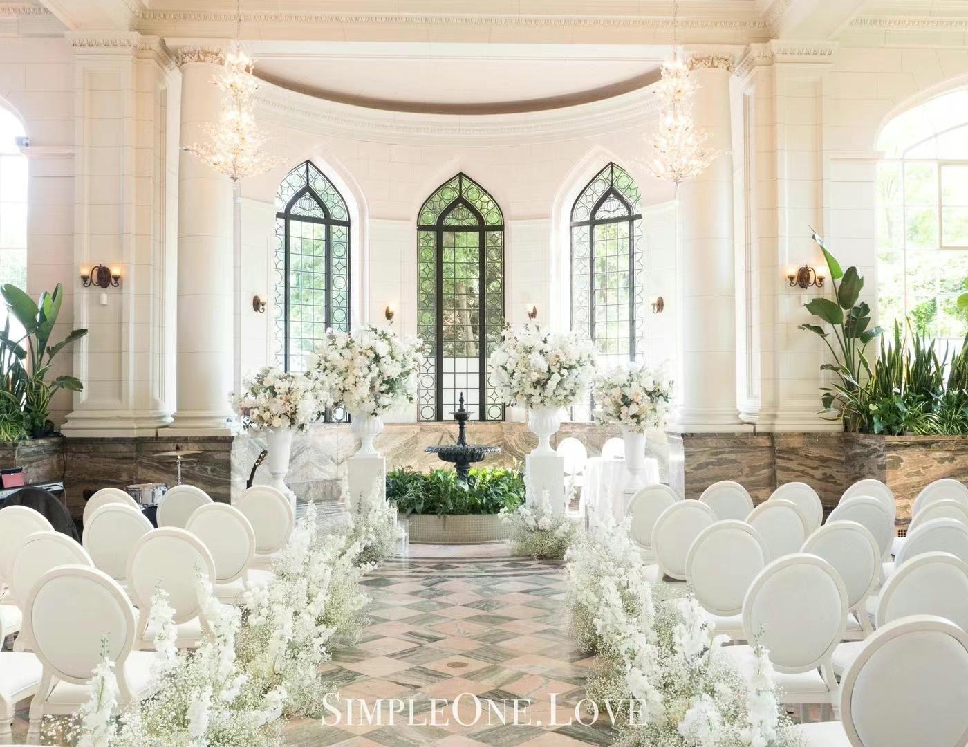 A conservatory ceremony: white chairs, arched windows and a fountain.